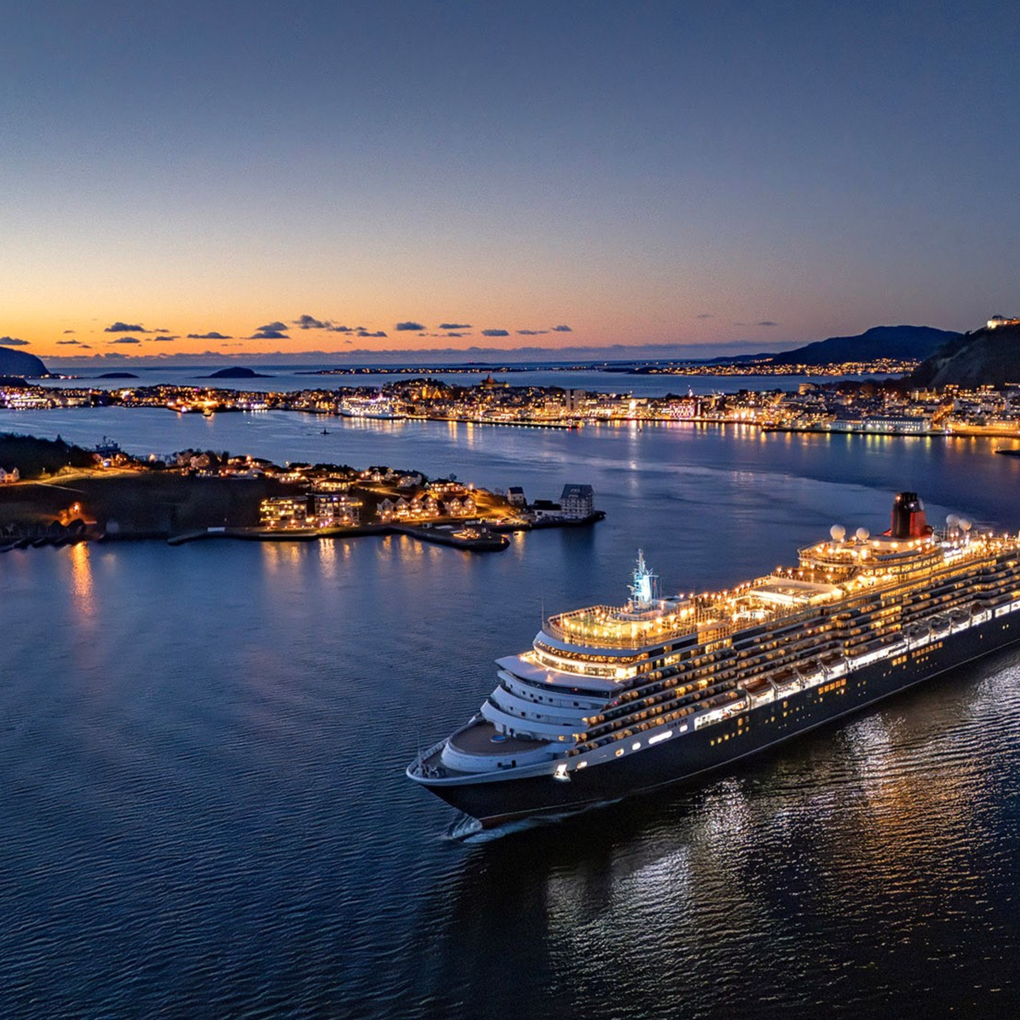 Eit cruiseskip seglar ut frå Ålesund i blåtimen – byen lysar opp i bakgrunnen